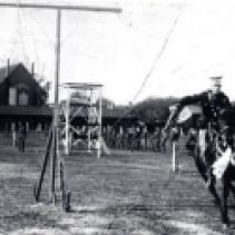T.L. Kennedy demonstrates the art of slicing a suspended lemon while galloping on horseback. He trained recruits at the Cooksville Fair Grounds in 1914 and the village is visible in the background.